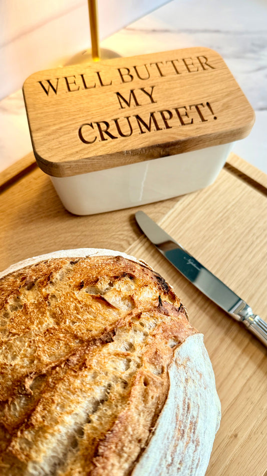 Personalised oak butter dish on a solid oak chopping board with fresh sourdough bread, a silver butter knife and soft natural lighting in a cosy kitchen.