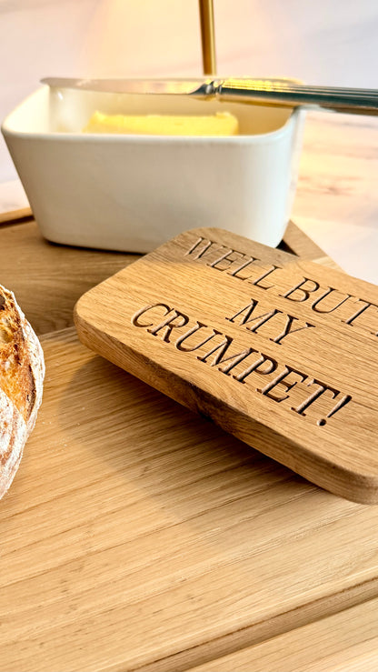 Wooden butter dish with 'Well but my crumpet!' text next to a butter dish on a wooden surface.