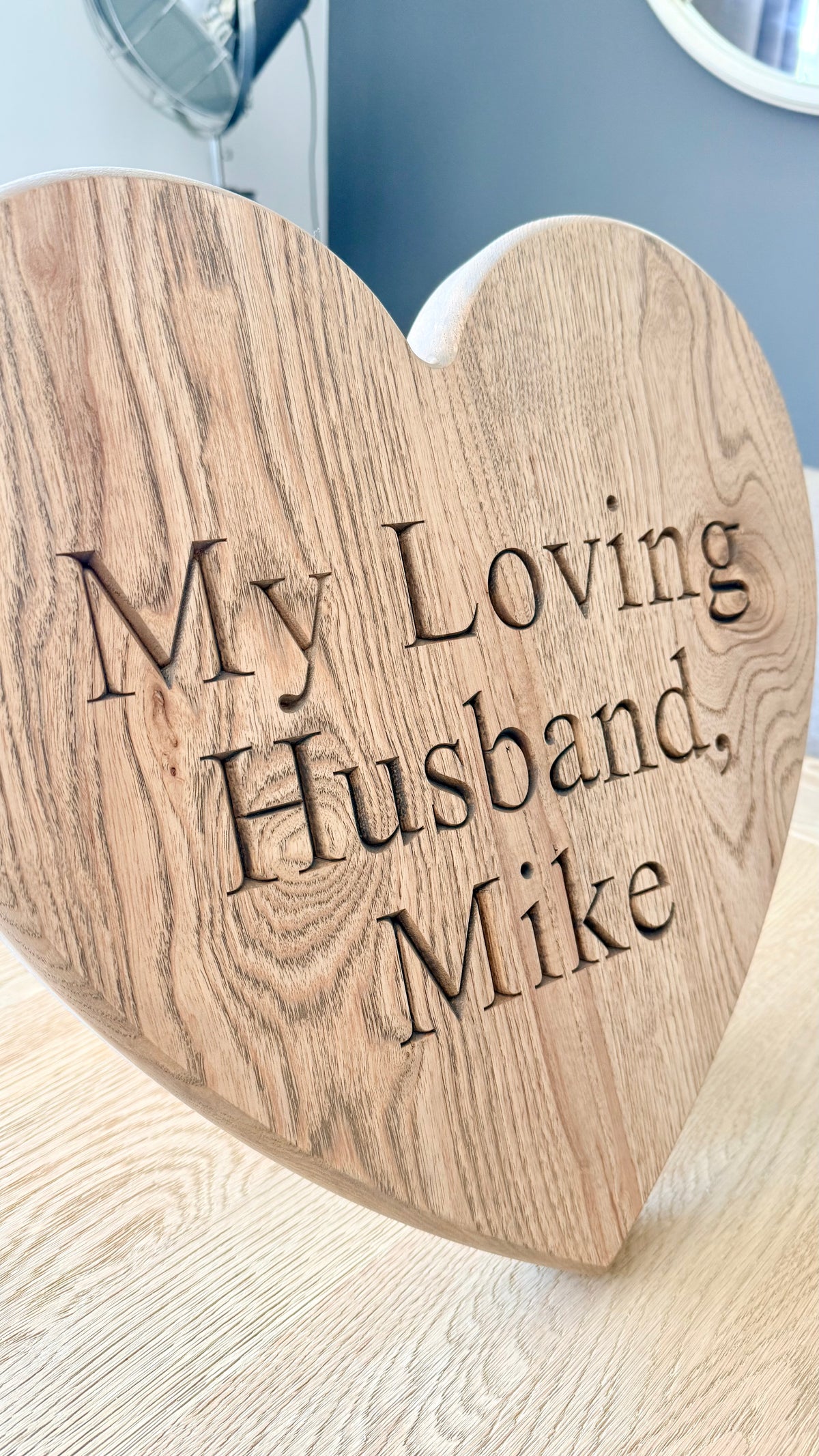 A heart-shaped wooden plaque engraved with the words 'My Loving Husband, Mike' in dark lettering, displayed on a light wood surface.