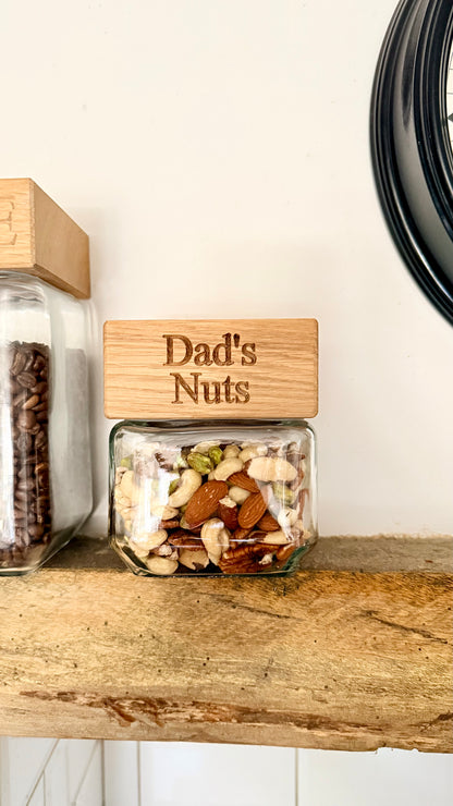 Jar labeled 'Dad's Nuts' on a wooden shelf with another jar in the background.