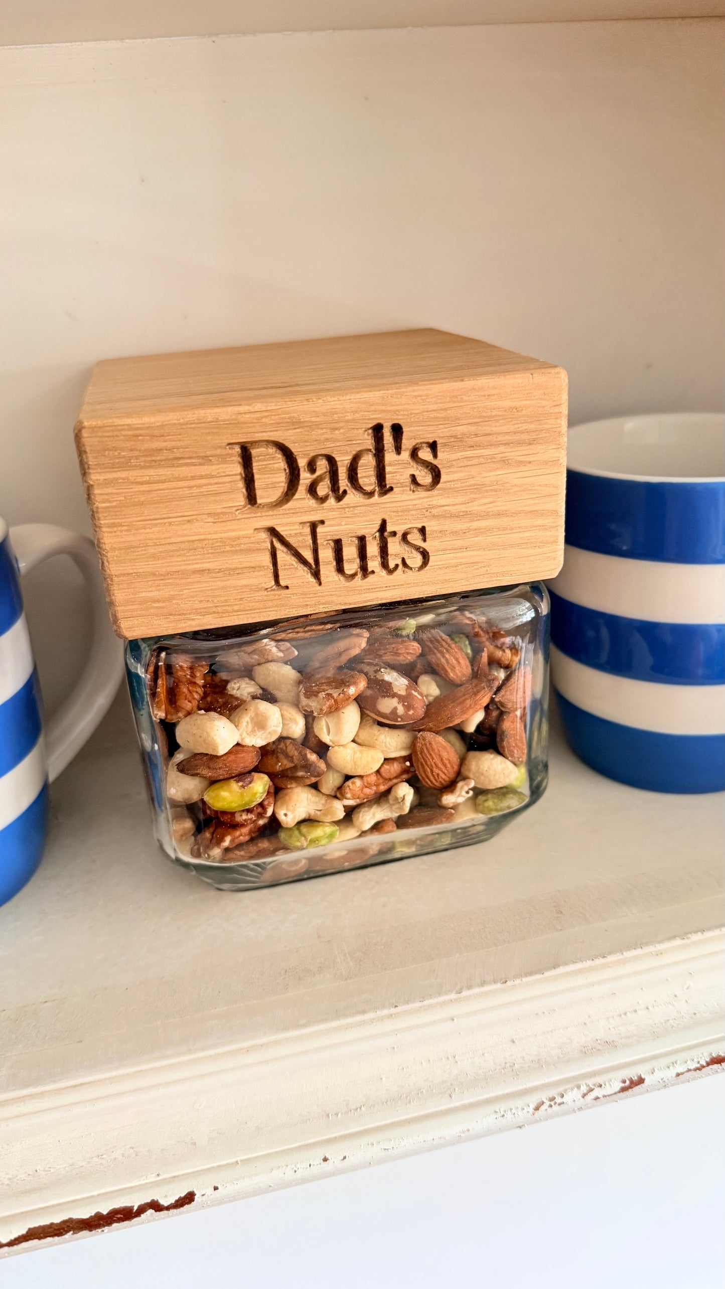 Glass jar with wooden lid labeled 'Dad's Nuts' on a shelf.