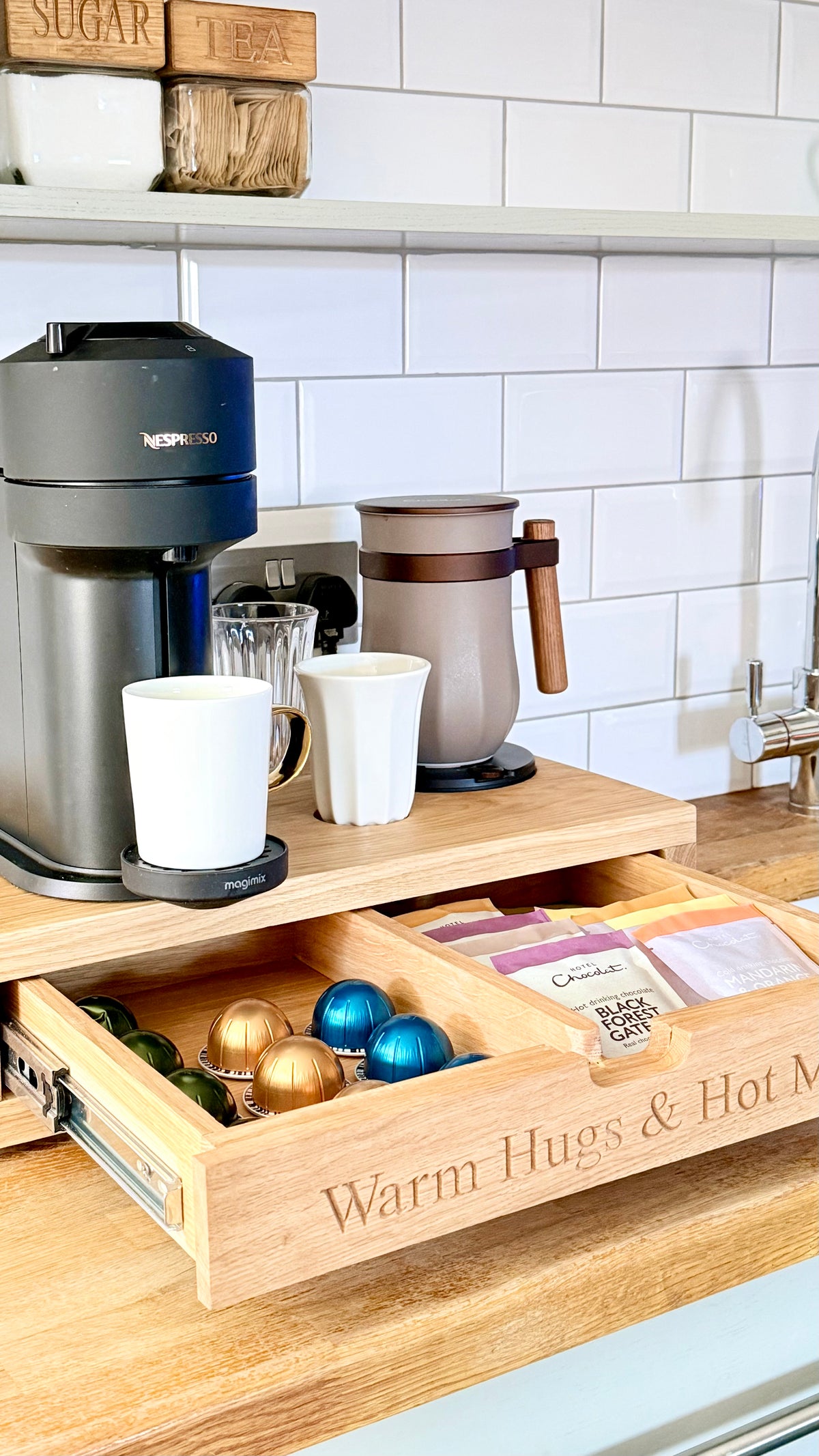 A wooden coffee station organizer on a kitchen counter, featuring a Nespresso coffee maker, a coffee pot, white mugs, and an open drawer labeled 'Warm Hugs & Hot M.