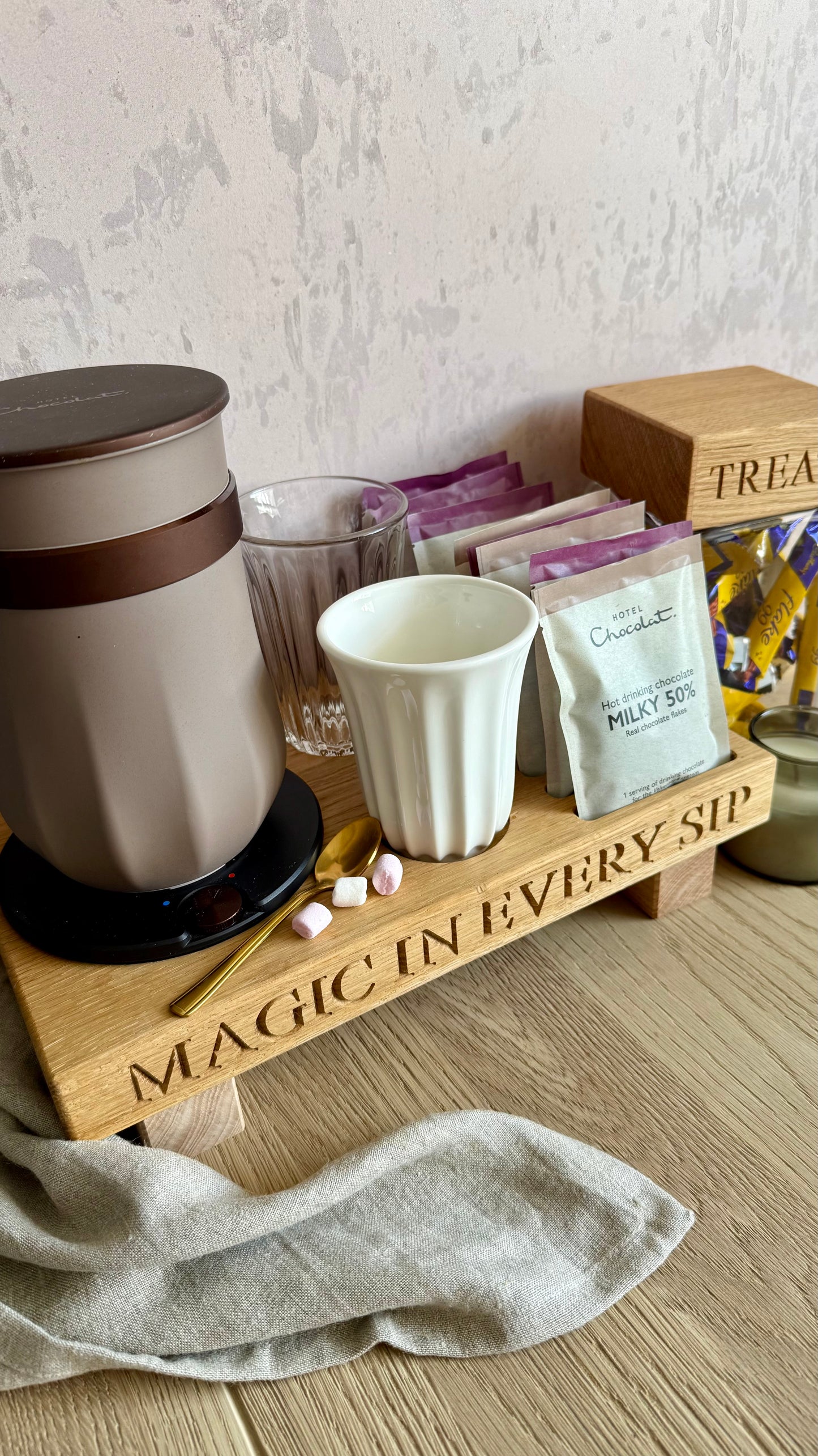 A wooden hot chocolate bar tray engraved with 'MAGIC IN EVERY SIP', holding a beige electric kettle, a white mug, chocolate and milk powder packets, a glass, marshmallows, and a candle, set on a wood floor against a textured wall.
