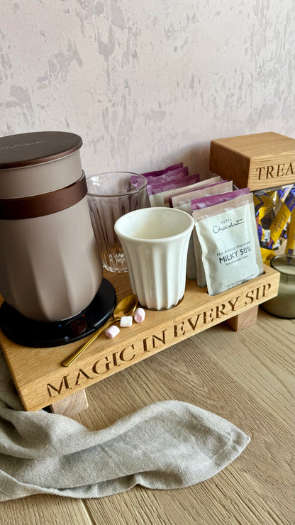 A wooden hot chocolate bar tray engraved with 'MAGIC IN EVERY SIP', holding a beige electric kettle, a white mug, chocolate and milk powder packets, a glass, marshmallows, and a candle, set on a wood floor against a textured wall.