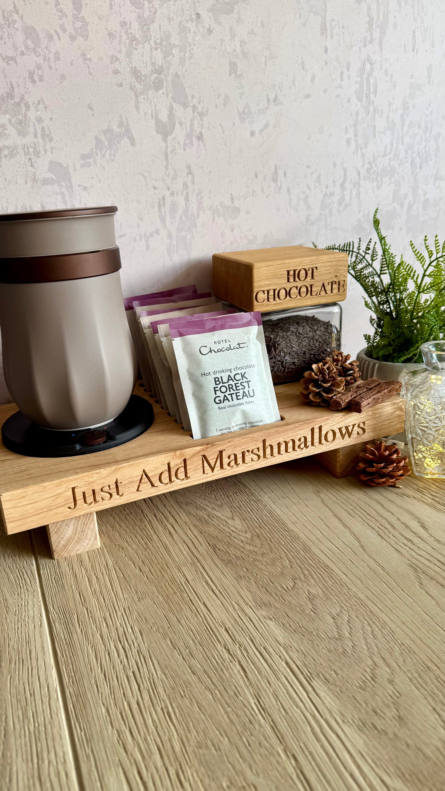 A wooden gift tray engraved with 'Just Add Marshmallows' holds assorted treats, including a mug, chocolate packets, a hot chocolate box, and a small plant, arranged on a light wood floor against a textured wall.