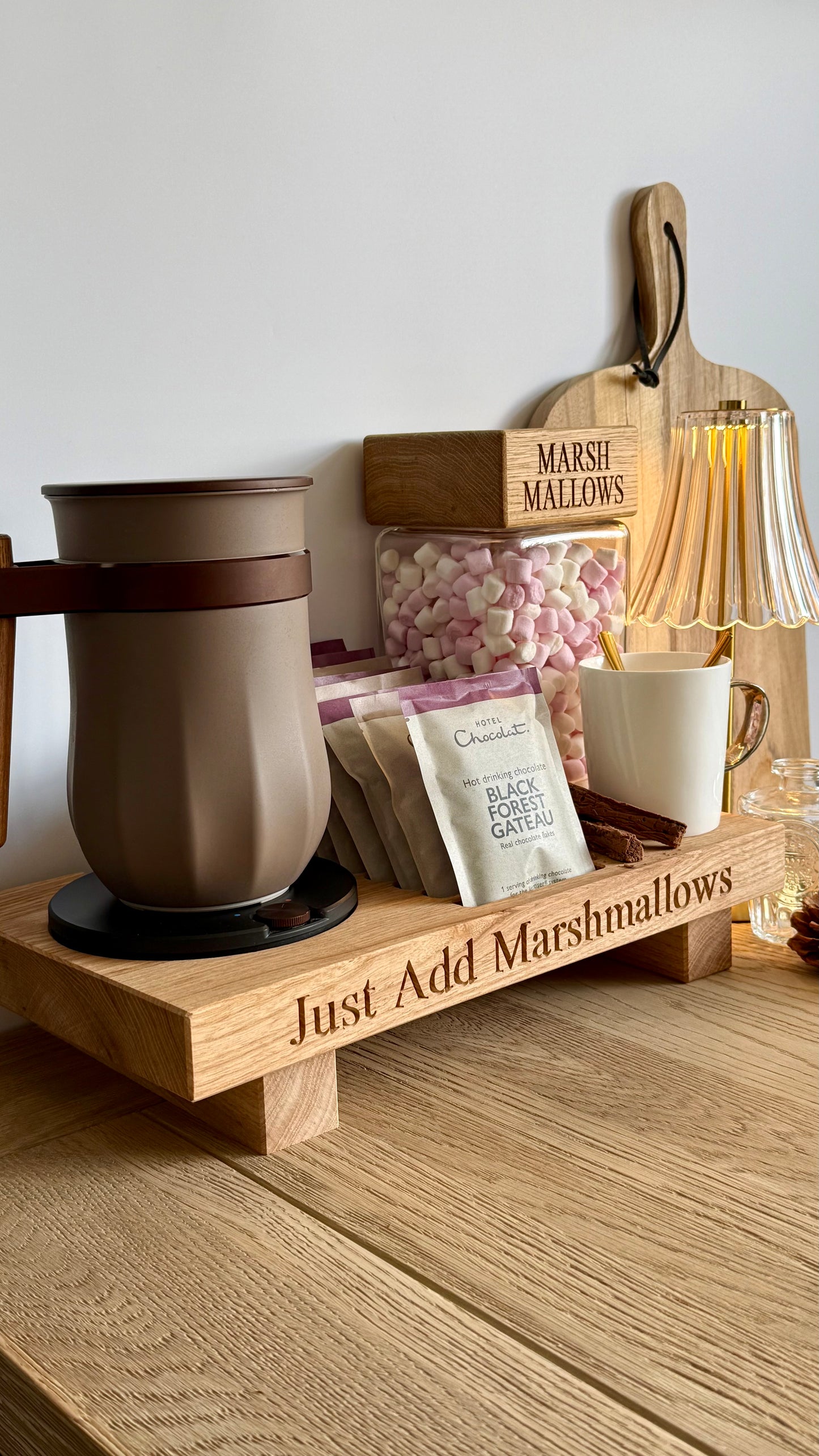 A wooden hot chocolate stationery tray engraved with 'Just Add Marshmallows' sits on a wooden table. The tray holds a brown insulated mug, a white mug with a spoon, packets of chocolate mix, a jar of pink and white marshmallows, and a wooden cutting board with 'Marshmallows' engraved on it.
