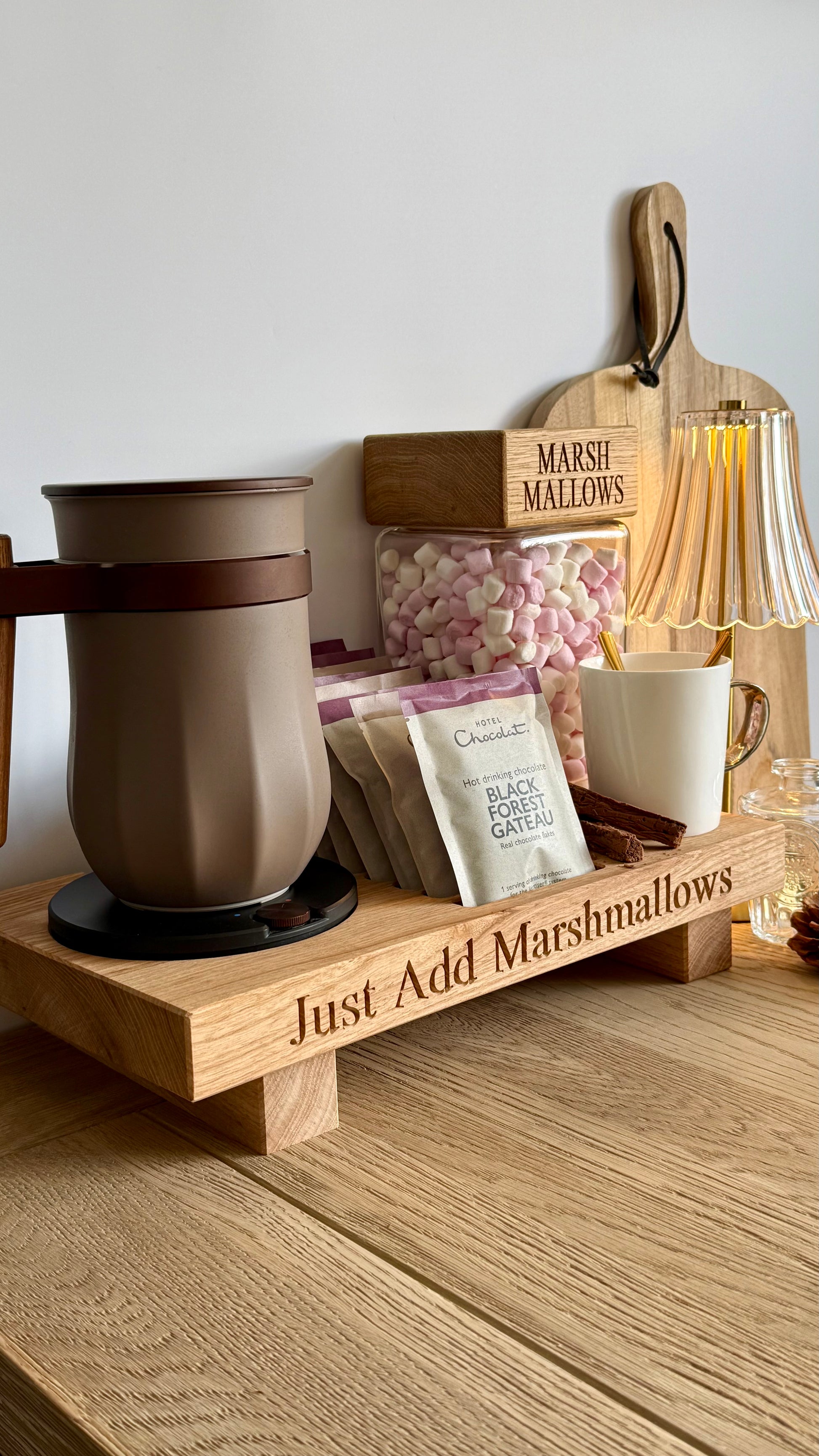A wooden hot chocolate stationery tray engraved with 'Just Add Marshmallows' sits on a wooden table. The tray holds a brown insulated mug, a white mug with a spoon, packets of chocolate mix, a jar of pink and white marshmallows, and a wooden cutting board with 'Marshmallows' engraved on it.