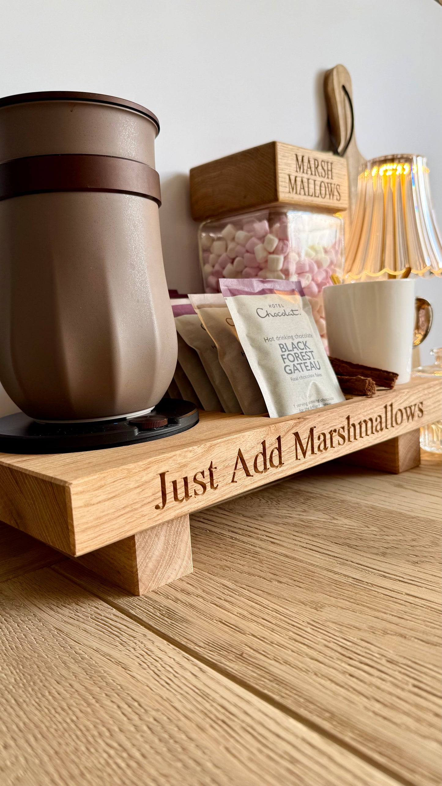 A wooden hot chocolate tray engraved with 'Just Add Marshmallows' sits on a wooden table. The tray holds a brown travel mug, a white mug, a bag of chocolate black forest garter mix, and a clear container of pink marshmallows.