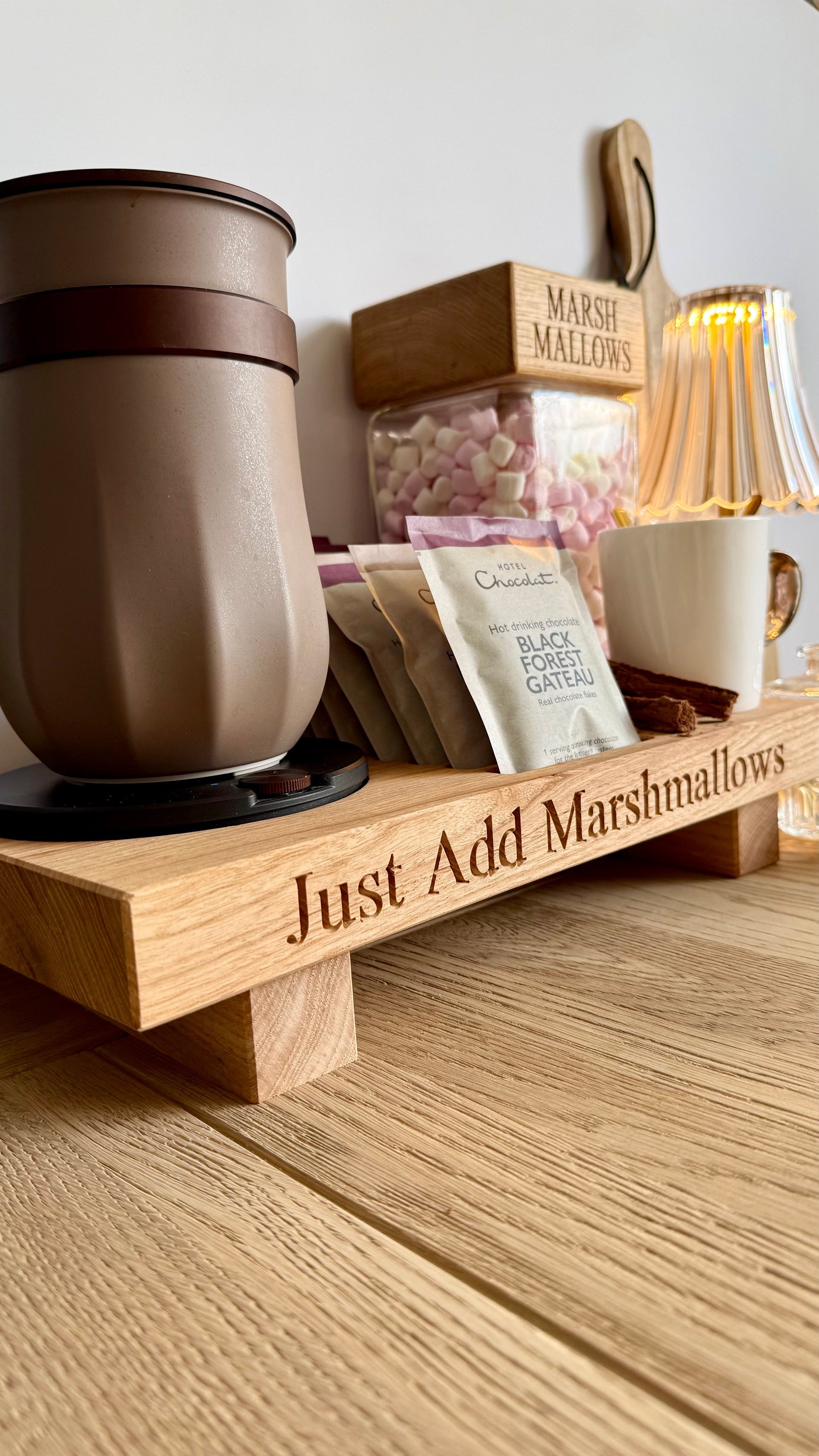 A wooden hot chocolate tray engraved with 'Just Add Marshmallows' sits on a wooden table. The tray holds a brown travel mug, a white mug, a bag of chocolate black forest garter mix, and a clear container of pink marshmallows.