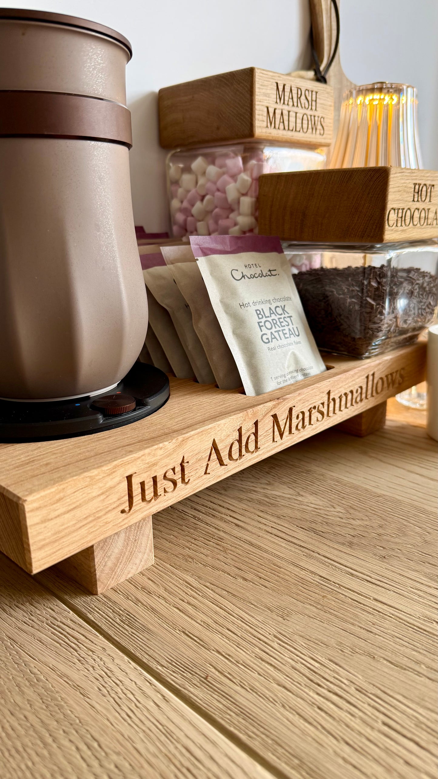 A wooden hot chocolate stationery set on a wooden tabletop, featuring a beige insulated mug, marshmallow jar, chocolate jar, and a wooden tray engraved with 'Just Add Marshmallows'.