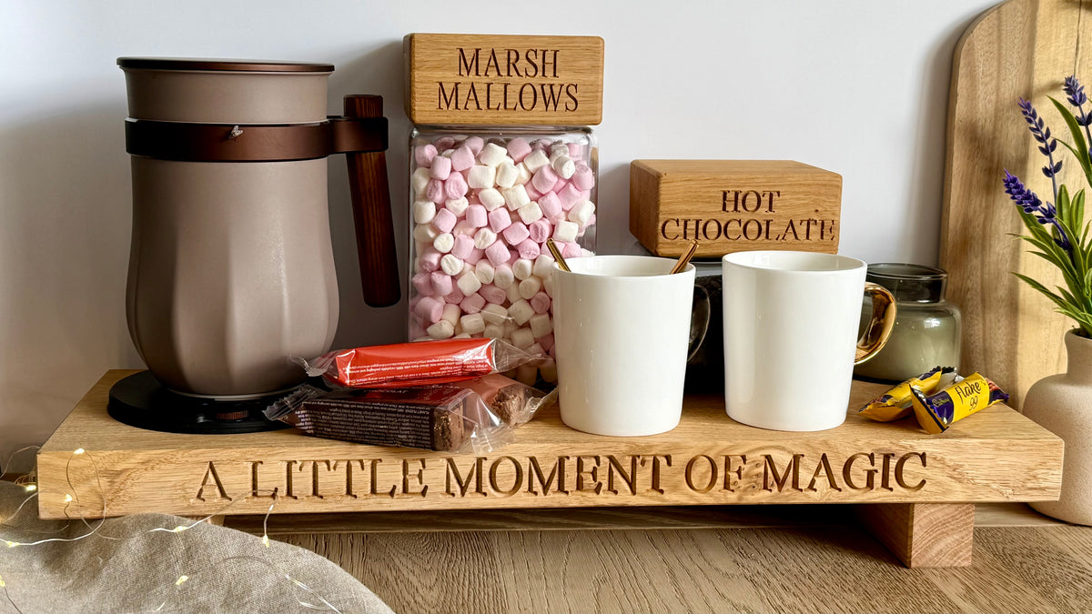 A hot chocolate bar setup on a wooden tray engraved with 'A LITTLE MOMENT OF MAGIC'. The tray holds a brown insulated mug, two white mugs, a box of marshmallows labeled 'MARSHMALLOWS', a box of hot chocolate labeled 'HOT CHOCOLATE', and chocolate bars.