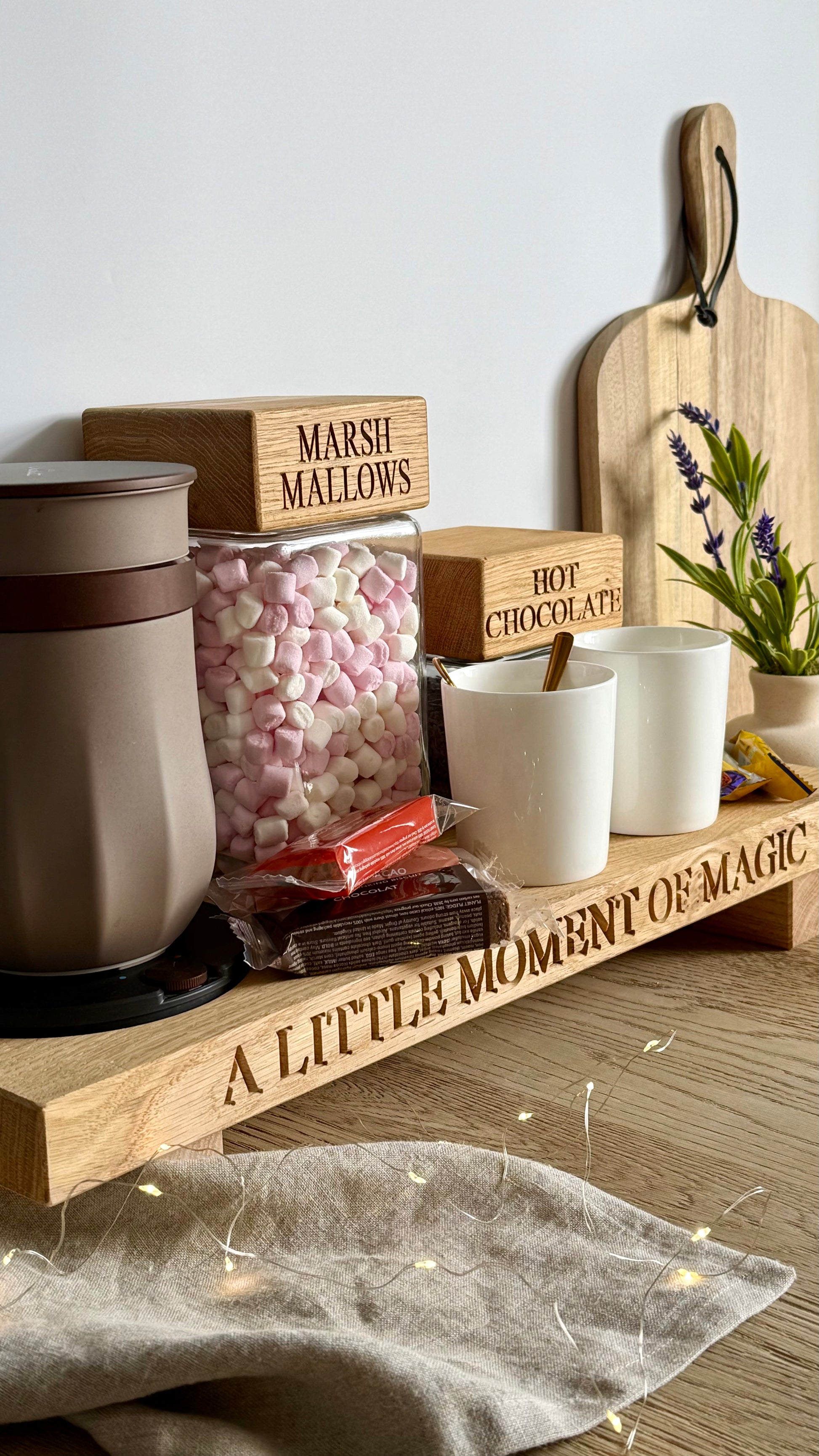 A styled hot chocolate stationery set on a wooden shelf, featuring a large insulated mug, a glass jar of marshmallows labeled 'MARSHMALLS', a box of hot chocolate labeled 'HOT CHOCOLATE', two white cups, and a wooden sign engraved with 'A LITTLE MOMENT OF MAGIC'.