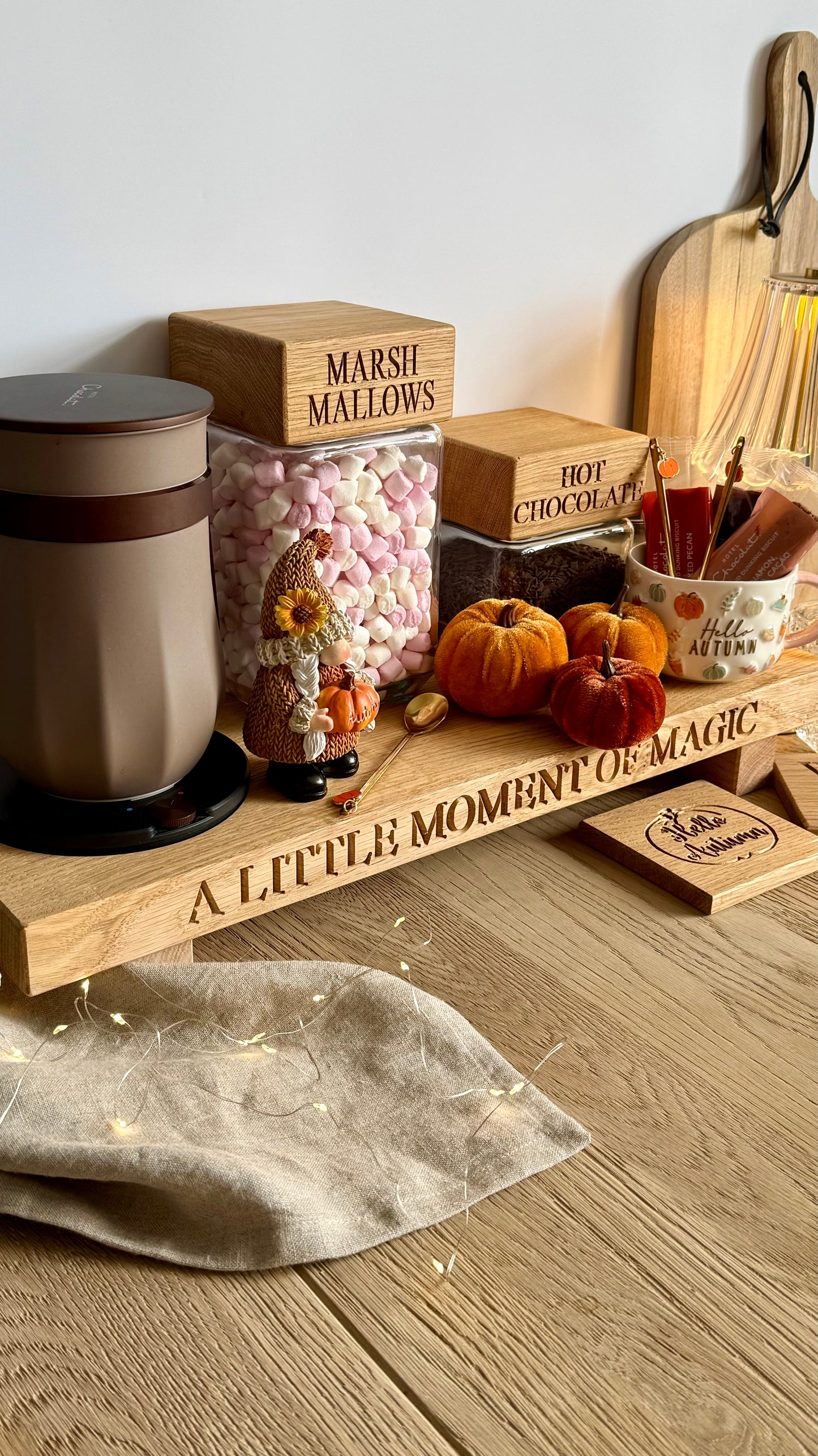 A styled tabletop arrangement in a warm kitchen or craft space, featuring a wooden tray engraved with 'A LITTLE MOMENT OF MAGIC,' clear jars labeled 'MARSHMALLOWS' and 'HOT CHOCOLATE,' small pumpkins, a mug with utensils, and string lights on a wood surface.