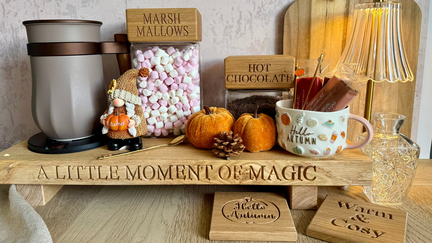 A styled autumn-themed wooden shelf display with a gray insulated mug on a base, a clear jar of pink marshmallows labeled 'MARSHMALLOWS,' a small decorative figure, a white mug with fall motifs and 'Autumn' text, two small pumpkins, pinecones, engraved wooden signs including 'A LITTLE MOMENT OF MAGIC,' and a warm glowing striped lamp in the background.