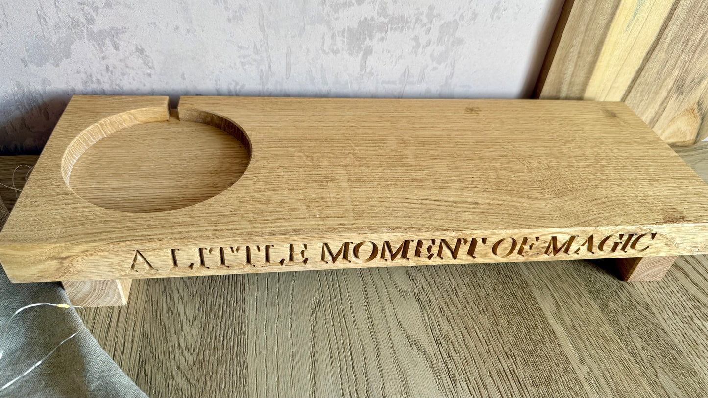 A rectangular wooden board with a circular cutout and engraved text reading 'A LITTLE MOMENT OF MAGIC', shown on a tabletop against a neutral indoor background.