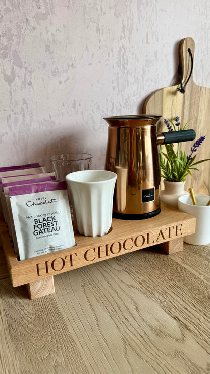 A wooden hot chocolate tray engraved with 'HOT CHOCOLATE' sits on a light wood table. The tray holds a white mug, a metallic copper milk frother, a bag of 'Cinq-Cents' black forest chocolate, and other chocolate packaging.