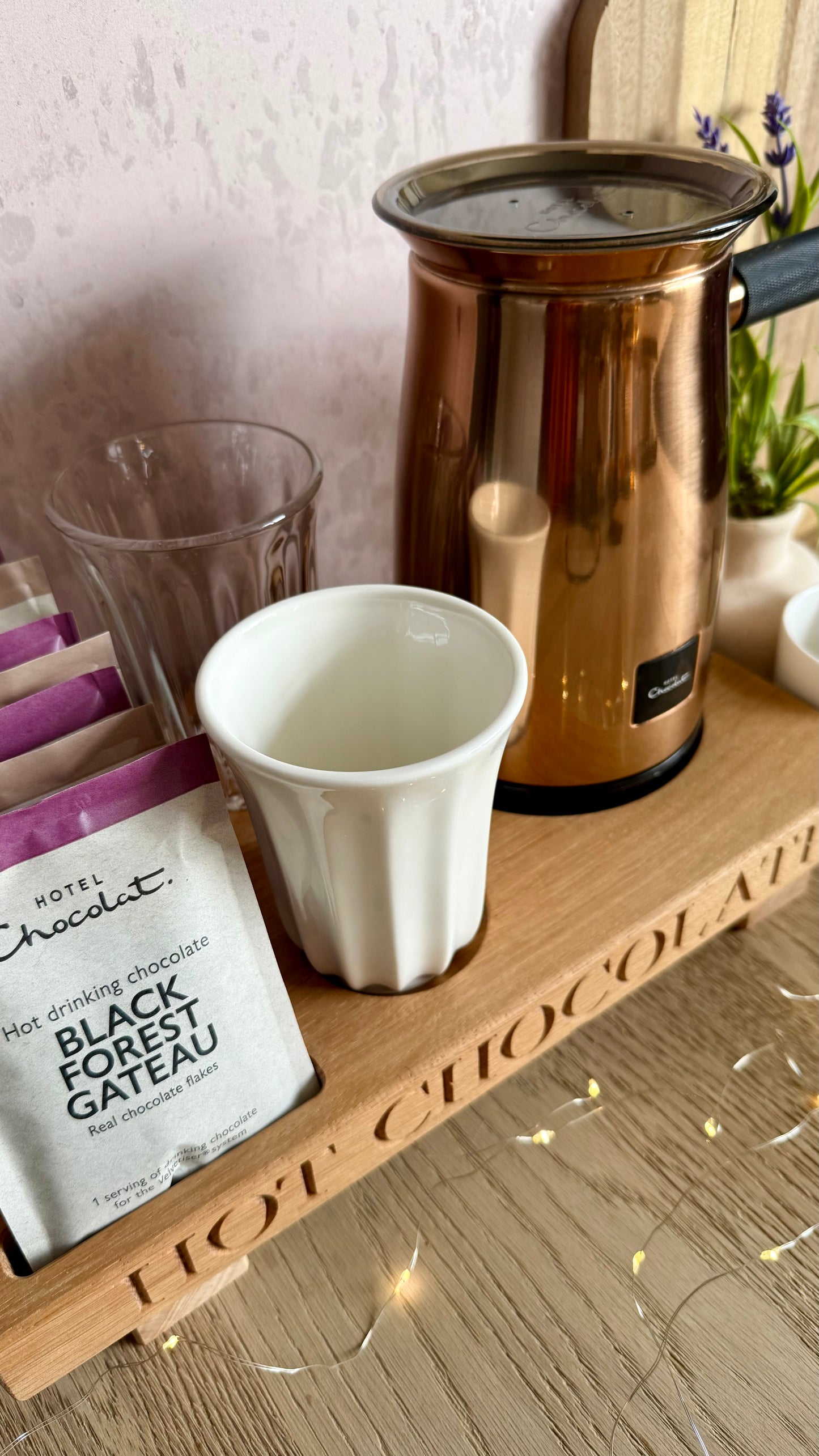 A styled hot chocolate setup on a wooden tray, featuring a white ceramic cup, a copper electric milk frother, a glass, and a bag of Hotel Chocolat Black Forest Gateau chocolate powder packets.