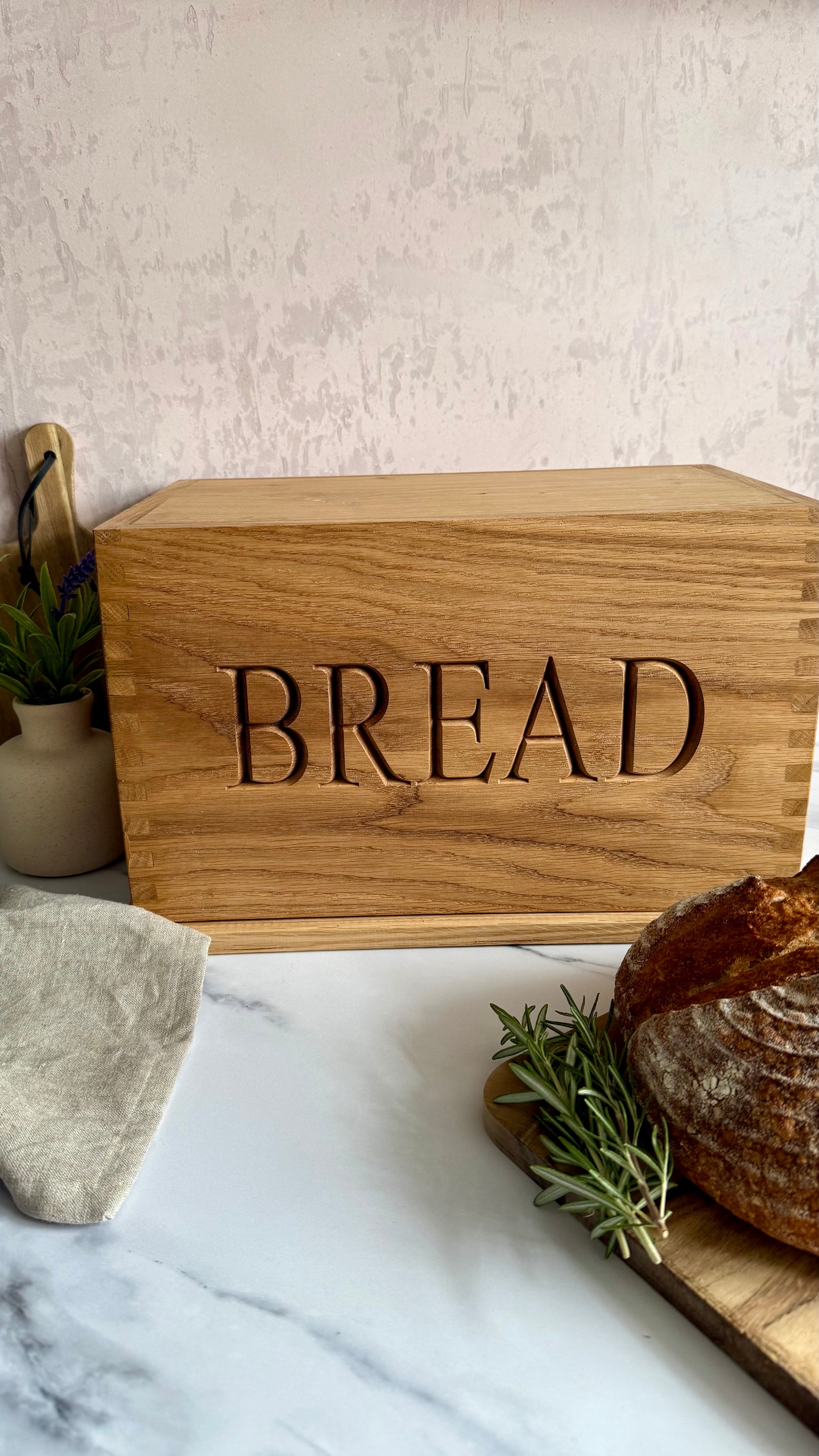 A rectangular wooden bread box engraved with the word 'BREAD' in large letters, styled on a white marble surface with a loaf of bread, rosemary, and a small plant nearby.