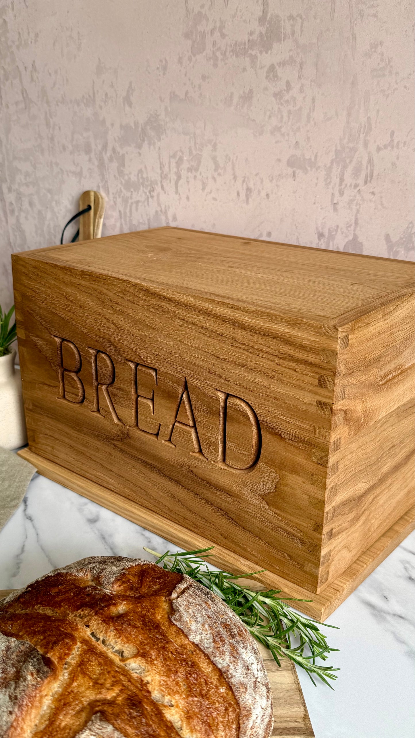 A rectangular wooden bread box engraved with the word 'BREAD' on the front, shown on a marble surface with a loaf of bread and rosemary in the foreground.