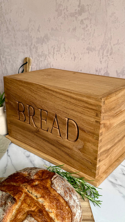 A rectangular wooden bread box engraved with the word 'BREAD' on the front, shown on a marble surface with a loaf of bread and rosemary in the foreground.