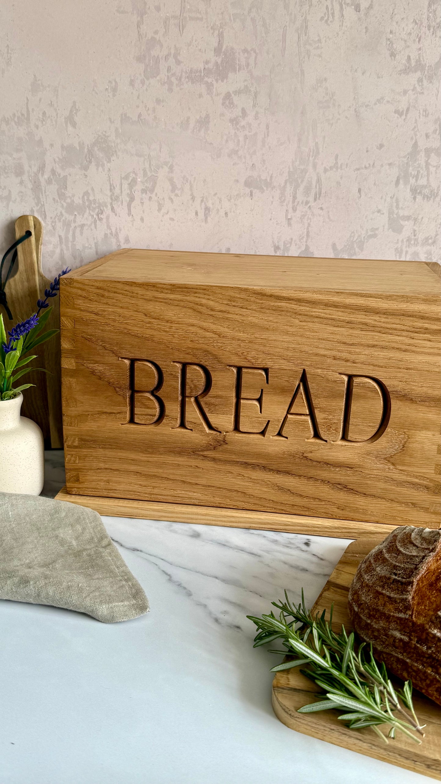 A rectangular wooden bread box engraved with the word 'BREAD' in large cutout letters, styled on a marble surface with a loaf of bread, rosemary, and a vase in the background.