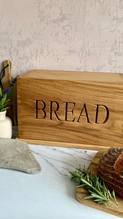 A rectangular wooden bread box engraved with the word 'BREAD' in large cutout letters, styled on a marble surface with a loaf of bread, rosemary, and a vase in the background.