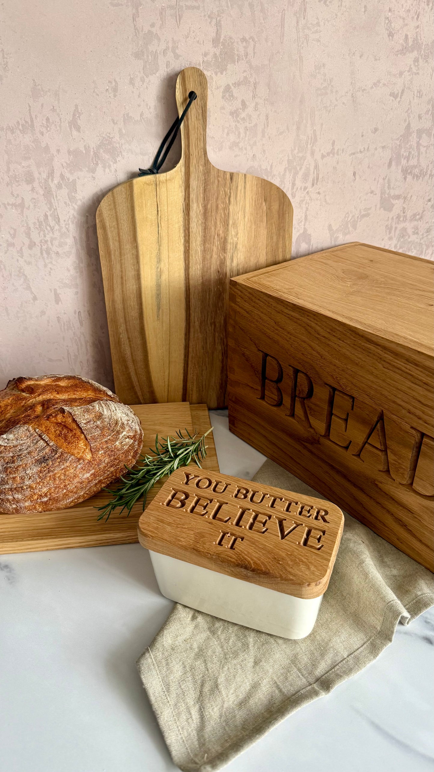 A styled kitchen scene with wooden cutting boards, a loaf of bread, a linen cloth, and a wooden butter box engraved with 'BREAD' and a smaller wooden container with 'YOU BUTTER BELIEVE IT' on the lid.