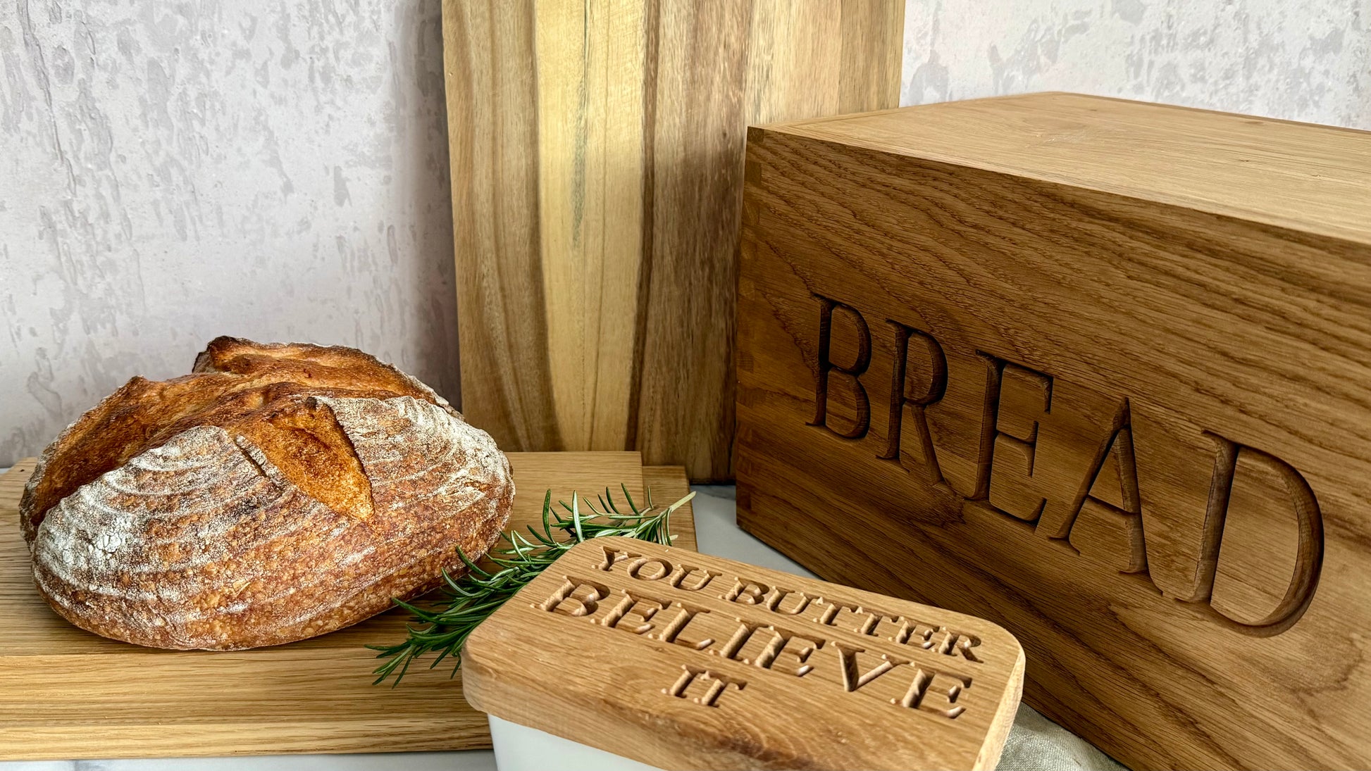 A rustic wooden bread box engraved with the word 'BREAD' stands next to a loaf of artisan sourdough bread on a wooden board, with a smaller wooden sign reading 'YOU BUTTER BELIEVE IT' in the foreground.
