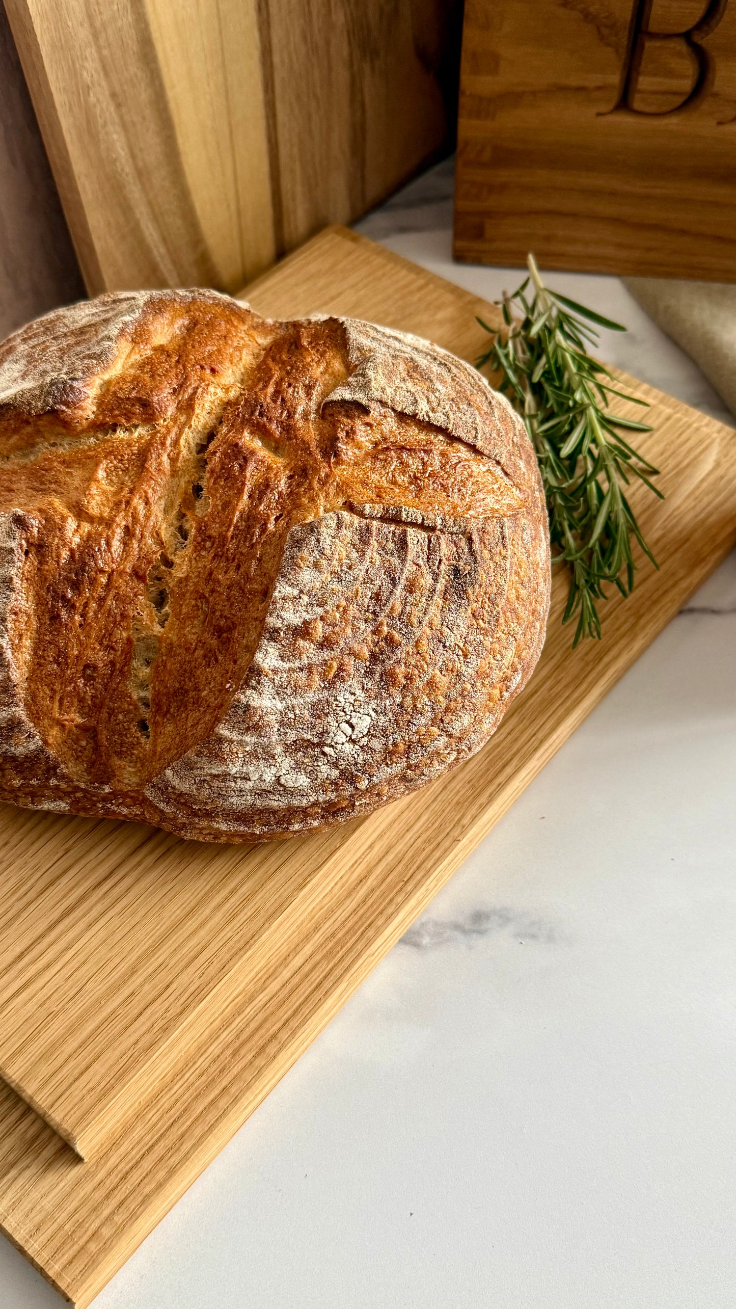 A round artisanal sourdough bread loaf with a golden-brown crust and flour-dusted surface, placed on a wooden cutting board with a sprig of rosemary beside it.