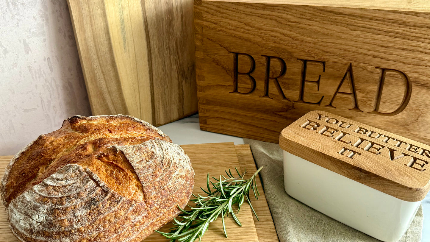 A rustic round loaf of bread on a wooden board next to a white butter dish with a wooden lid engraved with 'YOU BUTTER BELIEVE IT', with a wooden bread box in the background engraved with 'BREAD'.