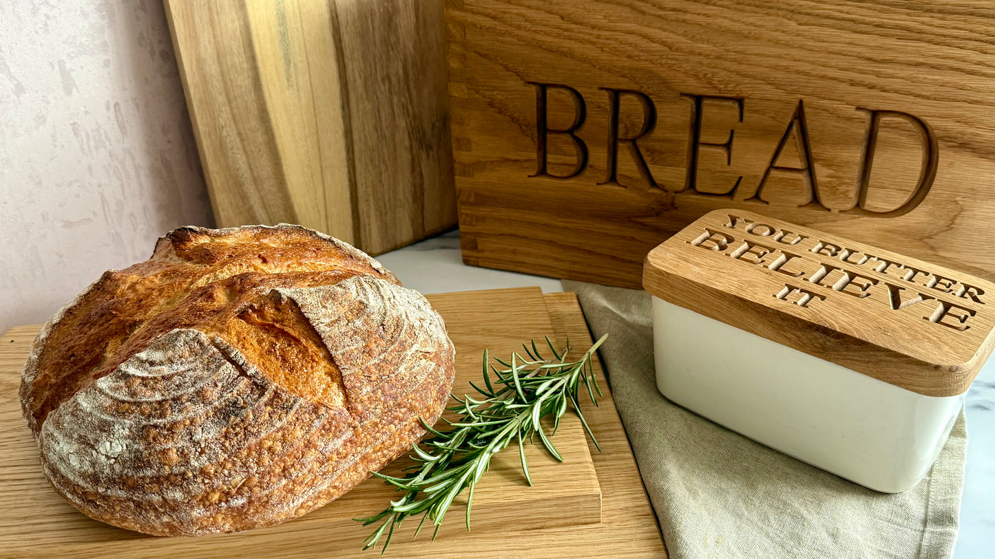 A rustic round loaf of bread on a wooden board next to a white butter dish with a wooden lid engraved with 'YOU BUTTER BELIEVE IT', with a wooden 'BREAD' box in the background.