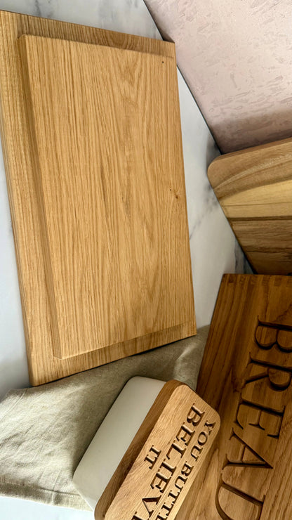 A set of wooden kitchen accessories including a large rectangular cutting board, a smaller wooden bread board engraved with 'BREAD', a wooden butter dish with 'YOU BUTTED IT BELIEVE IT', and a white butter container, all arranged on a marble surface.