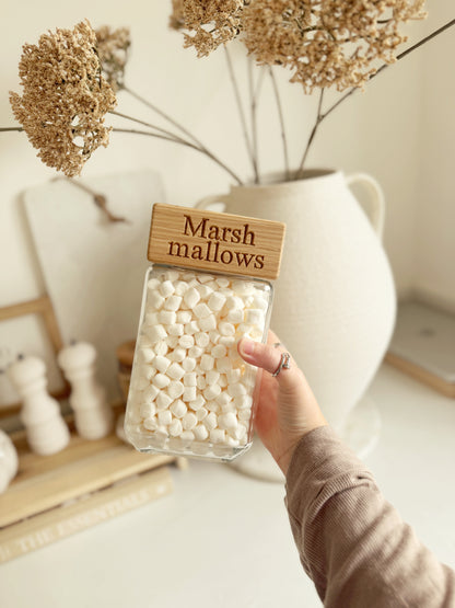 A hand holds a clear glass jar filled with mini marshmallows, topped with a wooden lid engraved with the words 'Marshmallows'. The background features a white ceramic vase with dried flowers and other neutral-toned home decor.