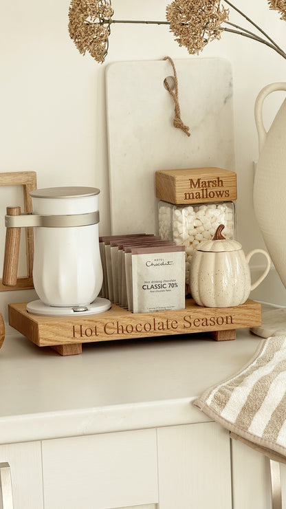 A wooden hot chocolate stationery tray on a white countertop, holding a white mug, a small cream pitcher, a jar of marshmallows, and a box of classic tea. The tray is engraved with 'Hot Chocolate Season'.